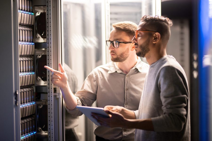 Team of specialists managing network server Team of young multi-ethnic specialists in glasses standing at server and using tablet while managing network server together in data center