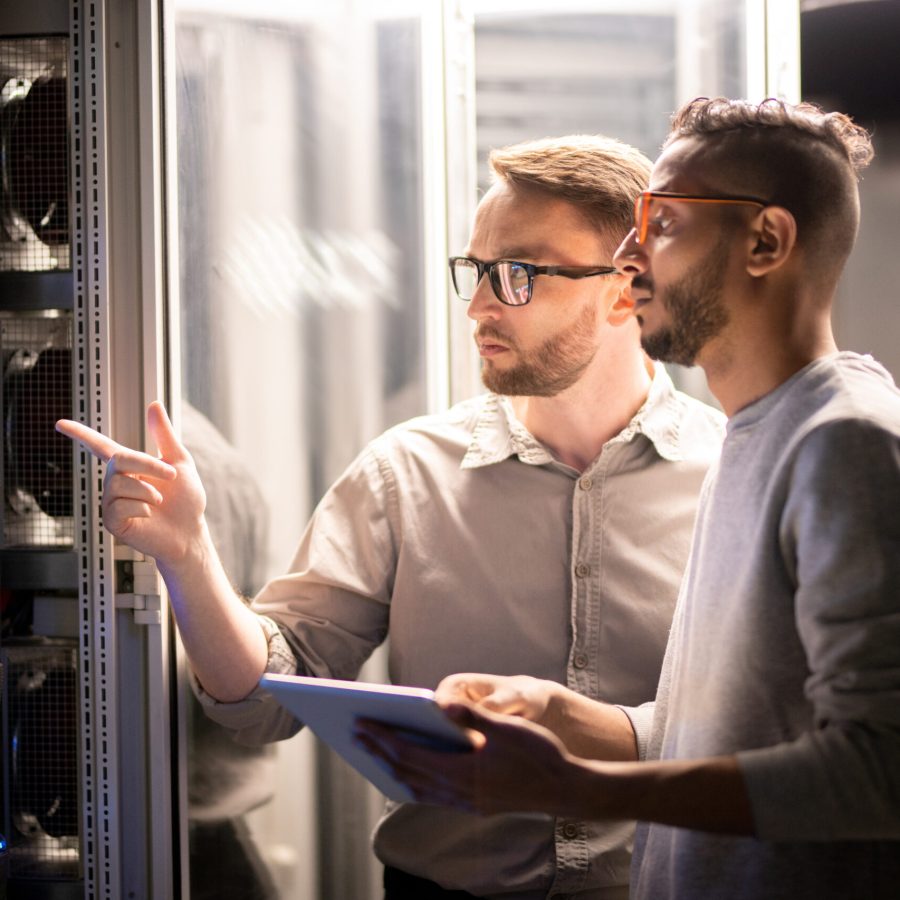 Team of young multi-ethnic specialists in glasses standing at server and using tablet while managing network server together in data center