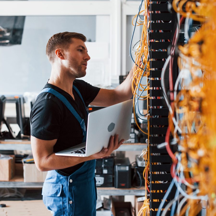 Young man in uniform and with laptop works with internet equipment and wires in server room Young man in uniform and with laptop works with internet equipment and wires in server room.