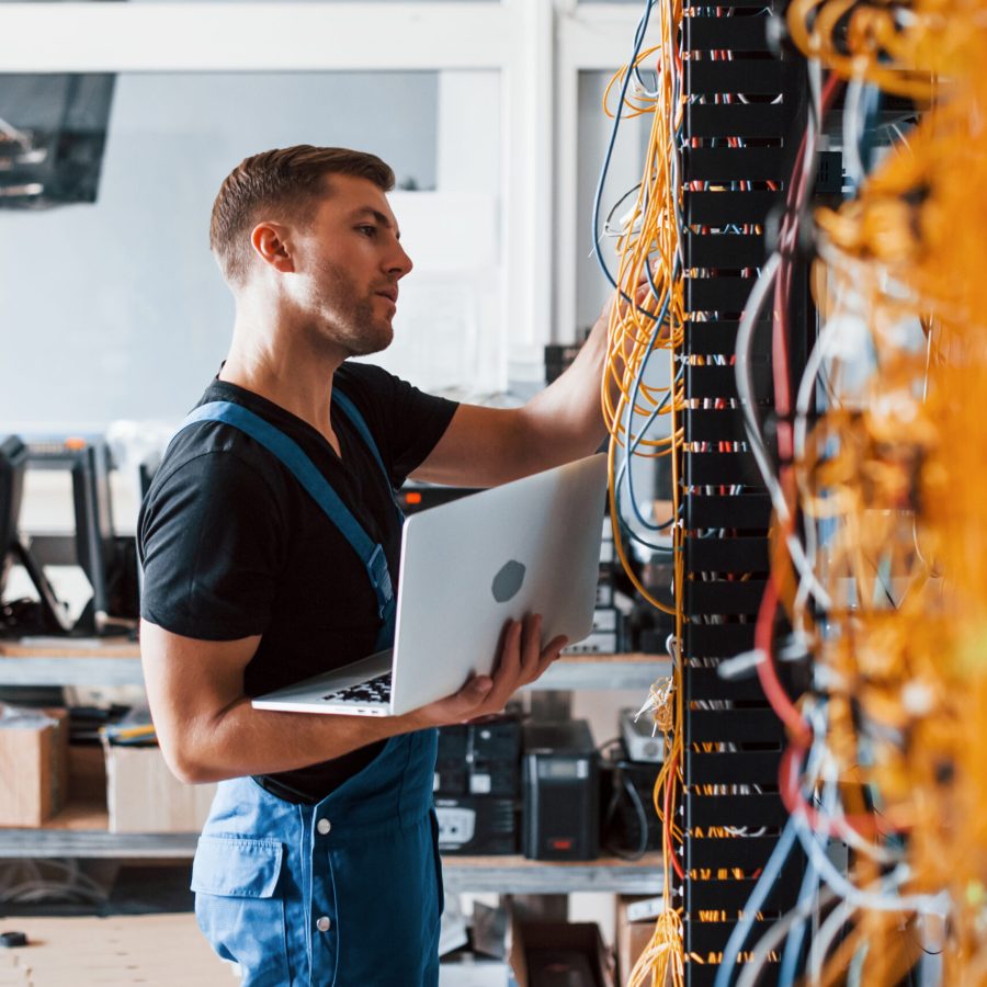 Young man in uniform and with laptop works with internet equipment and wires in server room.